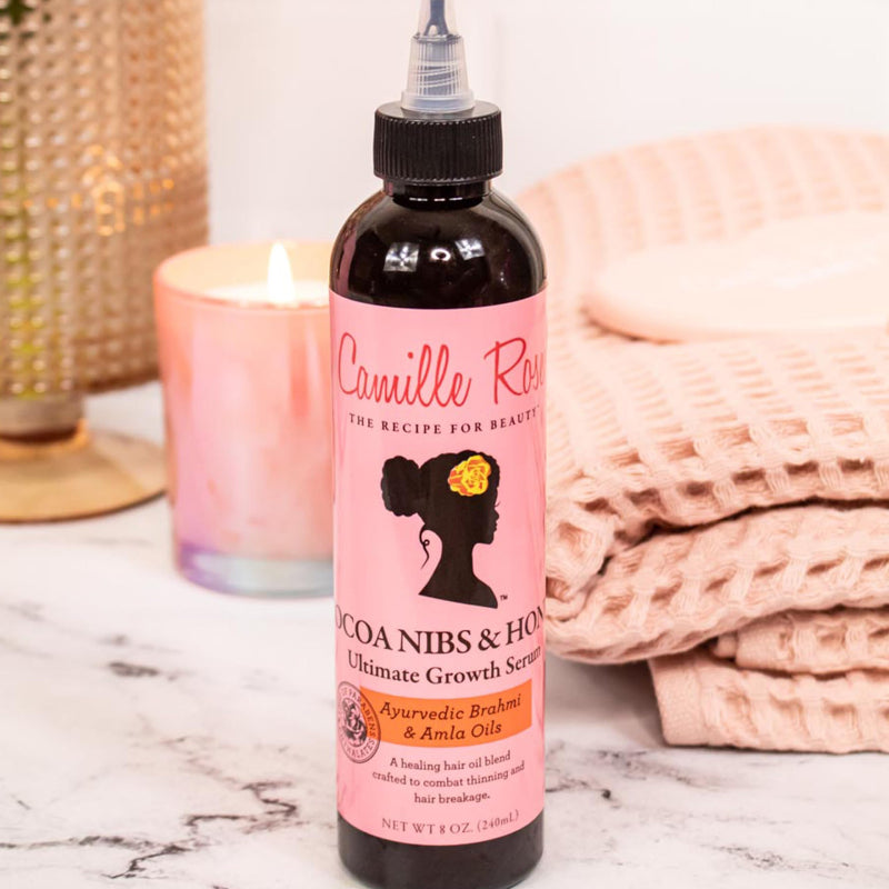 Camille Rose Cocoa Nibs & Honey growth serum bottle on a marble surface with a candle and blanket in the background.